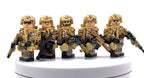 Five toy soldier minifigures in desert camouflage uniforms and helmets, each holding a black rifle, displayed on a white circular pedestal against a white background. Fabled Figs
