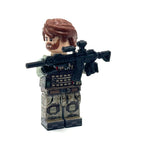 A custom-built toy minifigure with brown hair and beard, wearing a tactical vest and camouflage pants, holding a black rifle with a scope, set against a plain white background. Fabled Figs
