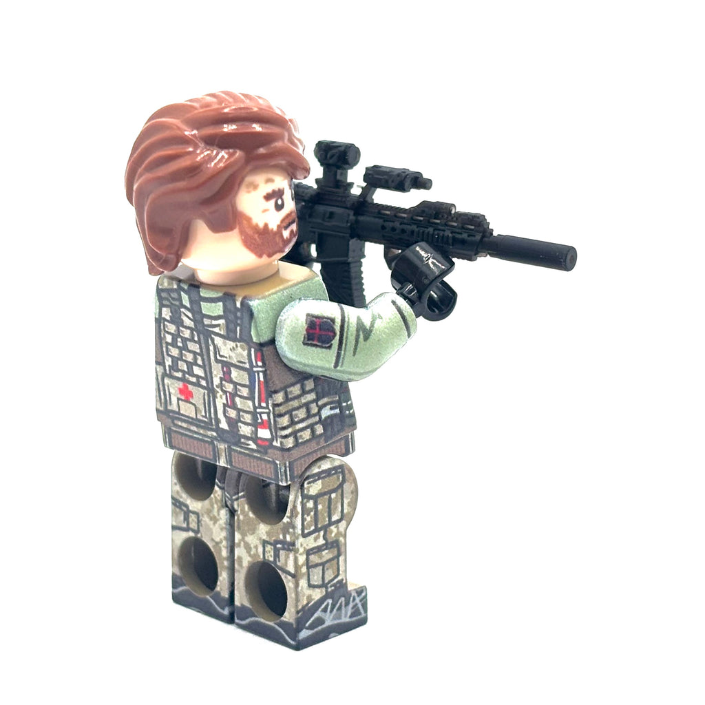 A custom-built toy minifigure with brown hair and beard, wearing a tactical vest and camouflage pants, holding a black rifle with a scope, set against a plain white background. Fabled Figs